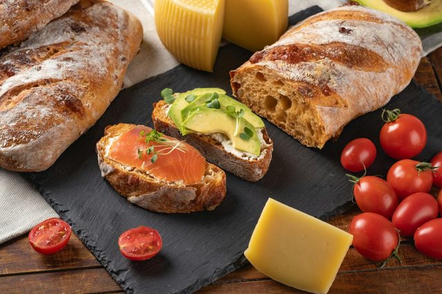 Sneden brood met beleg naast stokbrood, kaas en kerstomaatjes op een leisteen